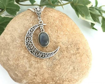 May include: A silver crescent moon pendant necklace with a dark blue labradorite gemstone. The necklace is displayed on a light brown rock.
