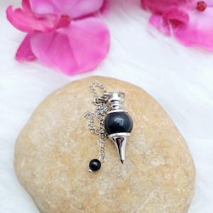 Könnte beinhalten: Ein schwarzes Obsidian-Pendel mit einer silbernen Kette und einer spitzen Spitze, das auf einem hellbraunen Stein liegt.