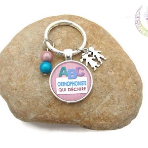 Könnte beinhalten: Ein rosa-blauer Schlüsselanhänger mit einem Anhänger, auf dem "Orthophoniste qui déchire" steht und die Buchstaben ABC darauf sind. Der Schlüsselanhänger hat auch zwei kleine Perlen, eine rosa und eine blaue.