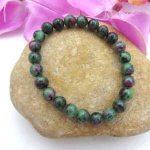 Op de afbeelding: Een groene en roze edelsteen armband met ronde kralen. De armband ligt op een lichtbruine steen.