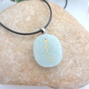 May include: A light green stone pendant necklace with a black cord. The stone has gold lettering that reads "本質", which is Japanese for "essence".