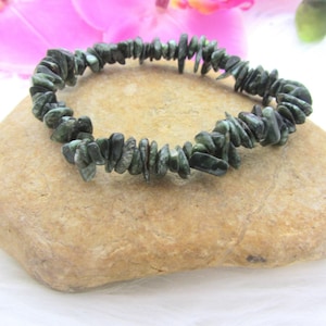 May include: A green and black gemstone chip bracelet made of natural stones. The bracelet is made of small, irregular shaped chips of green and black stone. The bracelet is on a light brown rock.