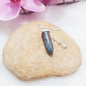 May include: A pendulum with a blue and brown stone, silver cap, and chain, with a clear crystal at the end. The pendulum rests on a beige stone, with a pink orchid in the background.