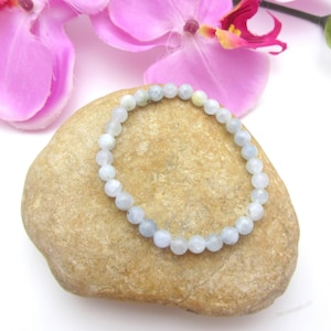 May include: A light blue beaded bracelet rests on a tan stone, with pink orchid blossoms in the background. The bracelet is made of small, round beads, likely gemstones, strung together to form a circular shape. The bracelet is a jewelry item.