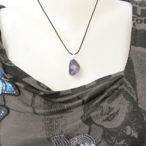 May include: A polished, irregular purple amethyst gemstone pendant suspended from a simple black cord necklace. The stone features natural variations in color and texture, hanging from a silver-toned metal bail against a dark patterned fabric.