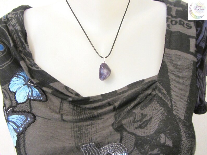May include: A polished, irregular purple amethyst gemstone pendant suspended from a simple black cord necklace. The stone features natural variations in color and texture, hanging from a silver-toned metal bail against a dark patterned fabric.