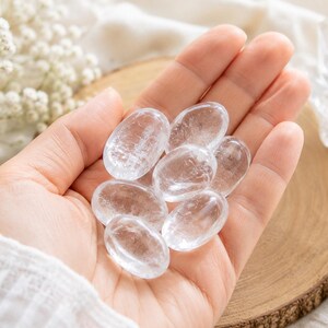 May include: A collection of six small, smooth, oval-shaped clear quartz crystals with a polished finish. Each stone is transparent with natural internal inclusions and fractures, reflecting light across their rounded surfaces while resting in a palm.