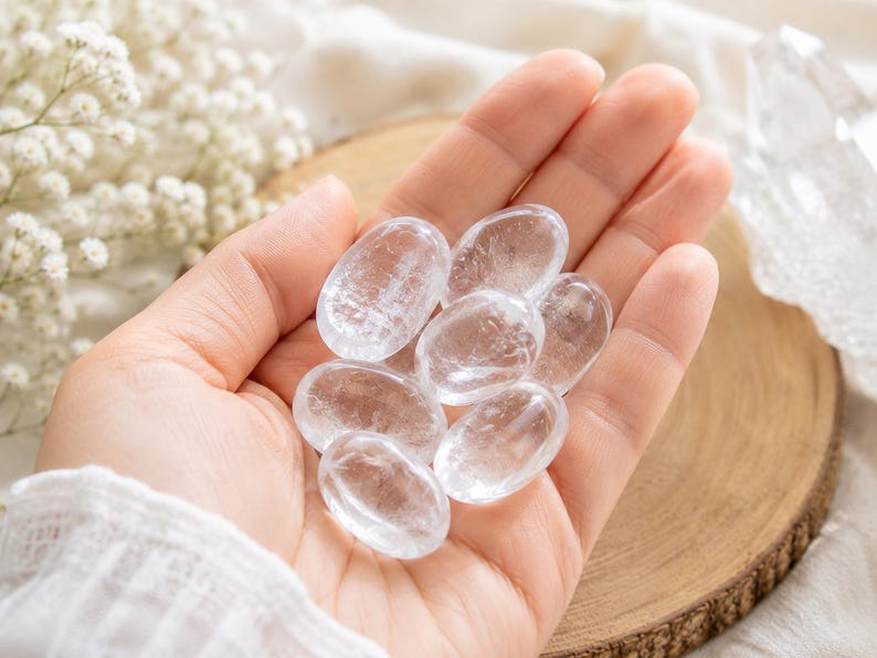 May include: A collection of six small, smooth, oval-shaped clear quartz crystals with a polished finish. Each stone is transparent with natural internal inclusions and fractures, reflecting light across their rounded surfaces while resting in a palm.