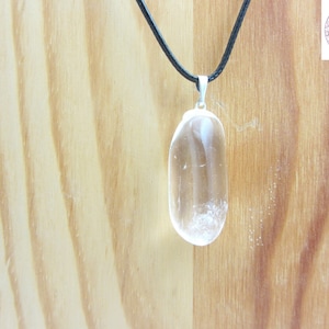 May include: A clear quartz crystal pendant on a black cord necklace. The pendant is oval shaped and has a smooth, polished finish.