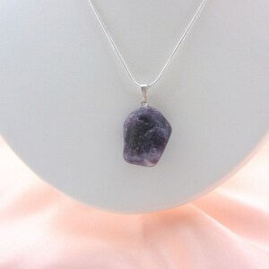 May include: A silver necklace featuring a pendant made of a rough-cut, dark purple gemstone. The pendant is suspended from a thin, silver chain. The necklace is displayed on a white, curved stand against a soft pink background.