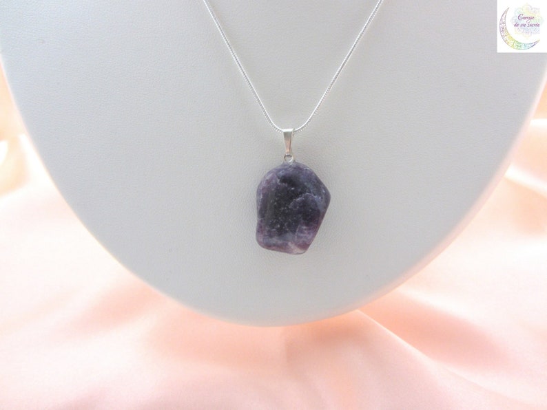 May include: A silver necklace featuring a pendant made of a rough-cut, dark purple gemstone. The pendant is suspended from a thin, silver chain. The necklace is displayed on a white, curved stand against a soft pink background.