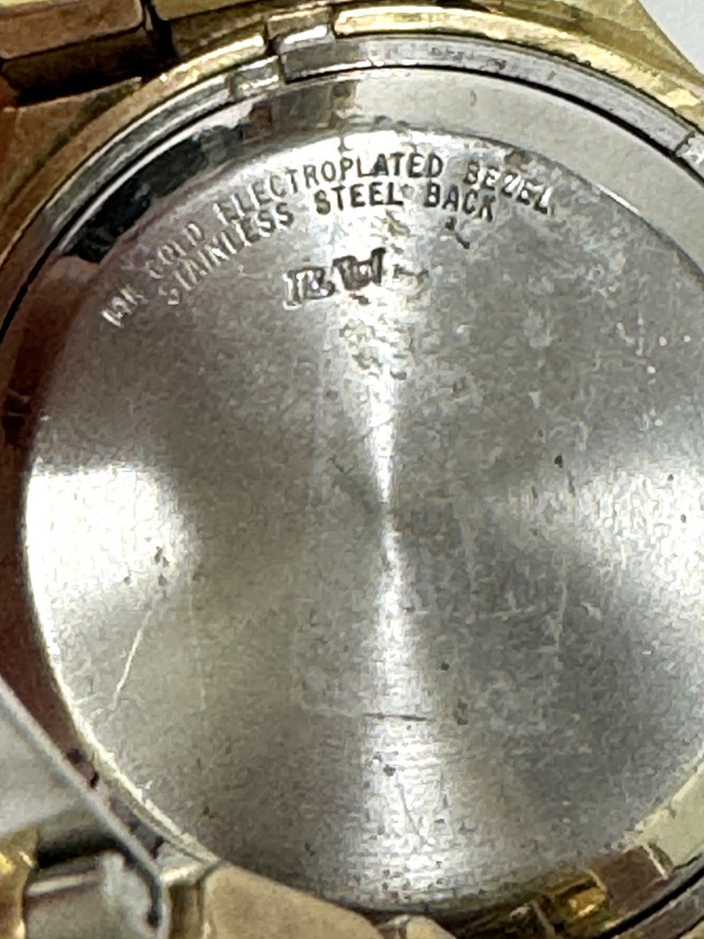 May include: Close-up of the back of a gold-plated watch with the text "14K Gold Electroplated Bezel Stainless Steel Back" engraved on the metal.