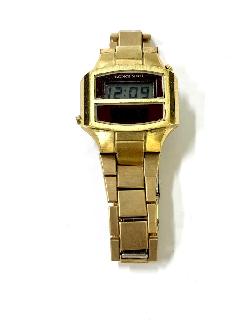 May include: A gold-toned Longines digital watch with a black digital display showing the time 12:09. The watch has a gold-toned metal band.