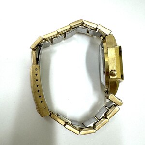 May include: A gold-tone wristwatch with a two-tone metal band. The watch has a square face and a gold-tone case.