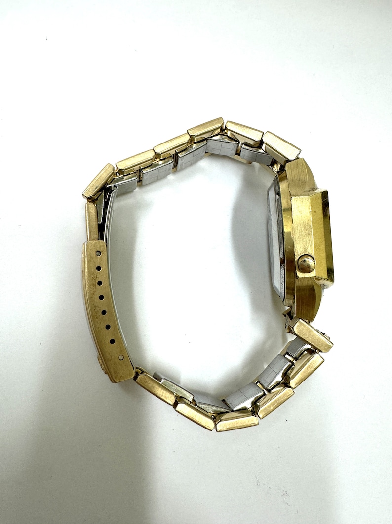 May include: A gold-tone wristwatch with a two-tone metal band. The watch has a square face and a gold-tone case.