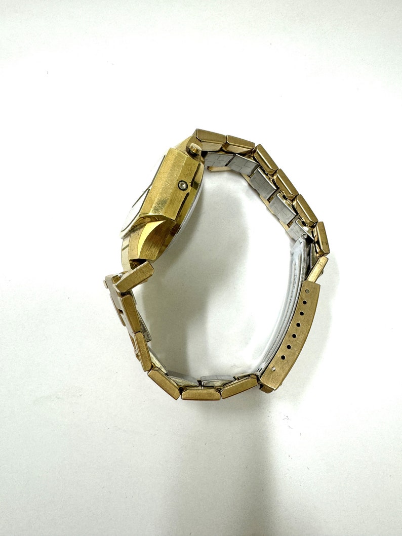 May include: A gold-toned wristwatch with a square face and a metal band. The watch is on a white background.