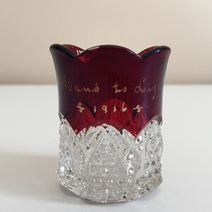May include: A small, clear glass cup with a red, scalloped rim. The cup has a cut glass pattern and is inscribed with "Mamie to Lizzie 1916".