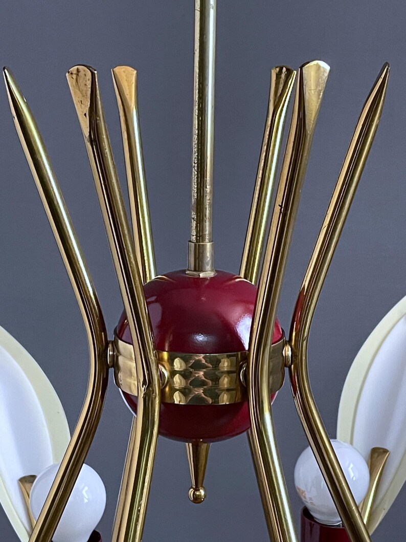 May include: A vintage chandelier featuring a central red sphere, brass-toned metal arms, and decorative details. The light fitting has white, teardrop-shaped shades and small, round light bulbs. The design is reminiscent of mid-century modern styles.