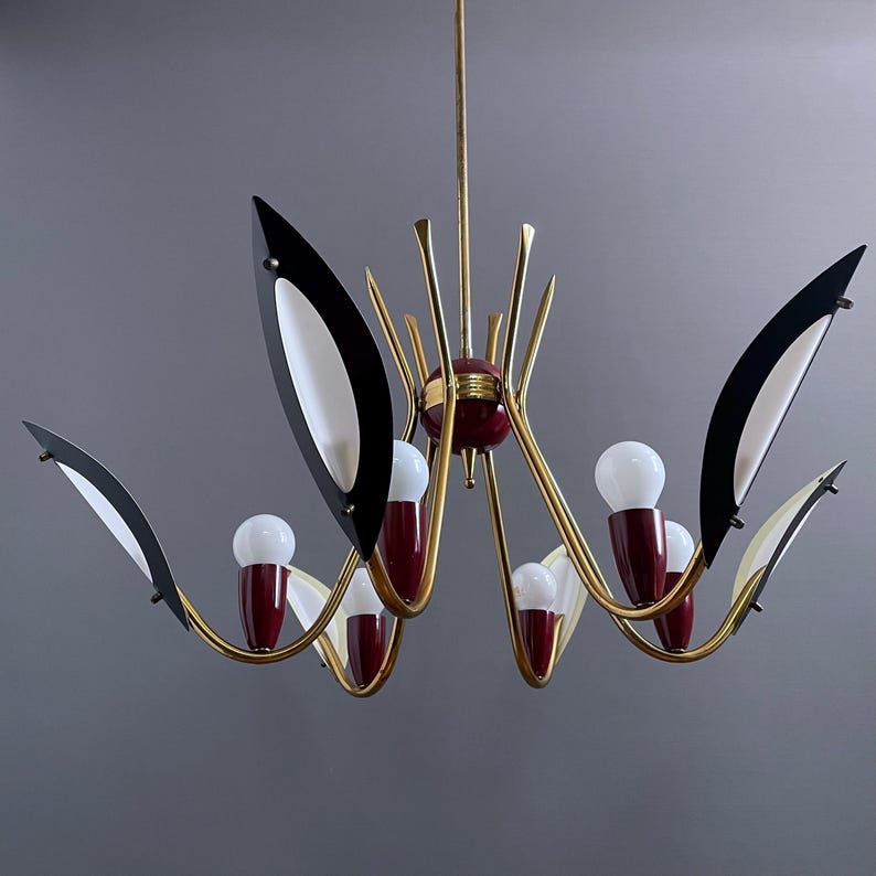 May include: A mid-century modern chandelier with a gold-tone frame, featuring black and white leaf-shaped accents. The chandelier has red and white bulb holders and multiple light bulbs, creating a stylish lighting fixture.
