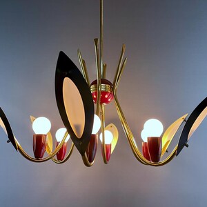 May include: A mid-century modern chandelier with a gold finish, featuring black and cream-coloured leaf-shaped accents. The chandelier has red accents and multiple light bulbs. A stylish lighting fixture for a home.