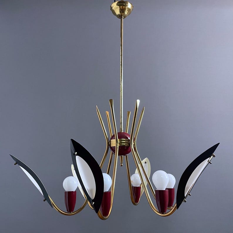 Può includere: Un lampadario moderno di metà secolo con finitura dorata, con accenti rossi e neri. L'apparecchio di illuminazione ha sei lampadine ed elementi decorativi a forma di foglia bianchi e neri. Il design è simmetrico ed elegante, con una sfera rossa centrale.