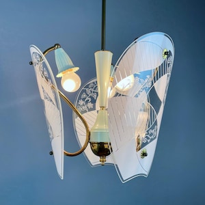 May include: A multi-arm pendant light fixture featuring three curved glass panels with white etched floral and geometric patterns. The central structure is cream-colored metal with brass accents, and it includes three light bulbs with light blue fluted covers.
