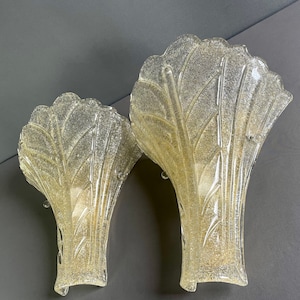 Puede incluir: Dos apliques de pared decorativos con diseño en forma de abanico. Los apliques son de vidrio transparente con efecto purpurina dorada. Los apliques tienen una superficie texturizada y están montados sobre un fondo gris. Para la decoración del hogar.
