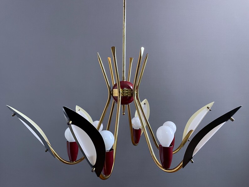 May include: A mid-century modern chandelier with a gold finish, featuring red and black accents. The light fixture has multiple arms with white bulbs and decorative elements, creating a stylish lighting design.