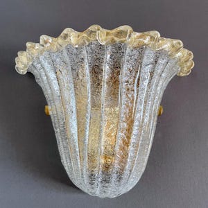 May include: A wall sconce featuring a clear, textured glass shade with a ruffled, gold-flecked rim. The shade has vertical fluted panels and is secured by two small, circular gold-toned metal fasteners on either side of the glass structure.