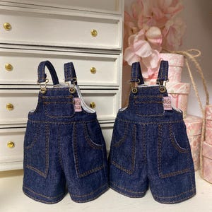 Puede incluir: Dos petos vaqueros en miniatura con costuras marrones y cierres metálicos. Los petos cortos tienen bolsillos delanteros y traseros y forro blanco. Los petos se exhiben sobre una superficie blanca con un armario blanco y flores rosas en el fondo.