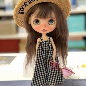 Op de afbeelding: Een pop met lang bruin haar, gekleed in een zwart-wit geruite jurk, zwarte schoenen en een strohoed met de tekst "some soul". De pop houdt een roze hartvormige zonnebril vast.