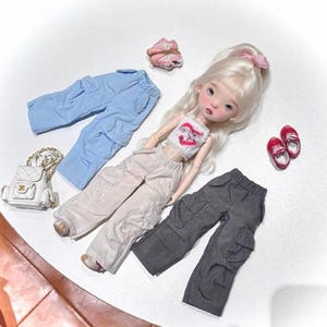 Può includere: Una bambola con lunghi capelli biondi e un fiocco rosa è circondata da vestiti per bambole. L'abbigliamento comprende pantaloni cargo azzurri, beige e grigi, un top corto bianco, scarpe da ginnastica rosa, scarpe da ginnastica rosse e una borsetta bianca.
