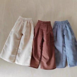 May include: Three pairs of wide-leg trousers in cream, rust, and light blue. The trousers have a high waist and pleats. The fabric looks like linen or a similar lightweight material, ideal for warmer weather. Each pair is a different shade.