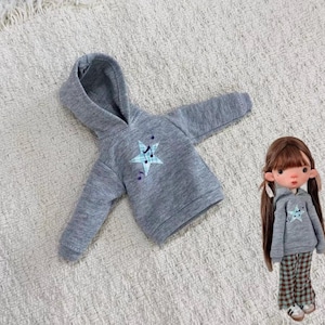 Può includere: Una felpa grigia a maniche lunghe con cappuccio caratterizzata da un'applicazione a forma di stella a quadri azzurro con accenti di note musicali blu scuro. Una bambola indossa una felpa grigia identica con pantaloni a quadri marroni e verdi.