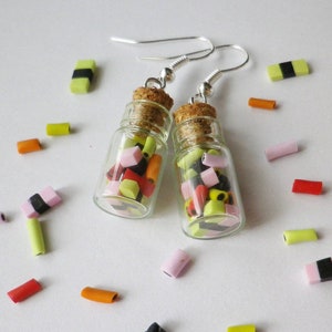 Pode incluir: Um par de brincos com pequenas garrafas de vidro cheias de doces coloridos. As garrafas são presas a ganchos prateados. Os doces são uma mistura de rosa, amarelo, laranja e preto.