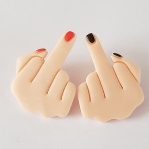 Può includere: Un paio di orecchini a forma di dito con un gesto di dito medio. Un orecchino ha lo smalto rosso e l'altro ha lo smalto nero.