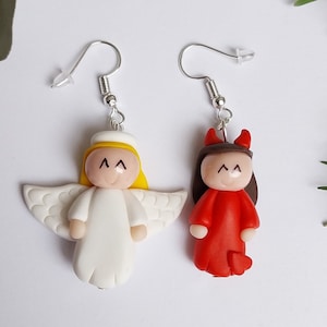 May include: A pair of dangle earrings featuring a white angel and a red devil. The angel has a yellow halo and the devil has horns. Both figures have black eyes and a smiling expression.