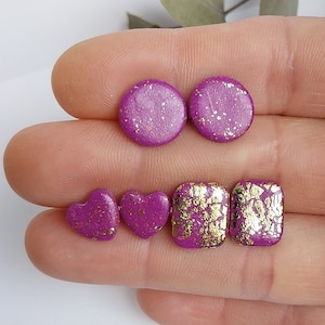 Peut inclure: Ensemble de six boucles d'oreilles clous violettes et dorées. Deux boucles d'oreilles rondes avec une finition scintillante, deux boucles d'oreilles en forme de cœur et deux boucles d'oreilles carrées avec des accents de feuille d'or.