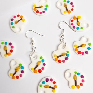 May include: A pair of silver earrings with white paint palette charms. Each charm has a small paintbrush and colourful dots.