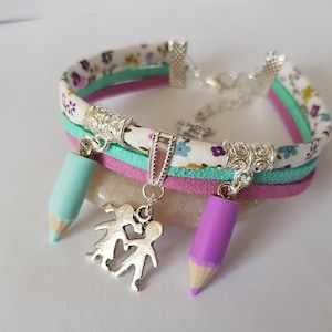 May include: A multi-layered bracelet with a floral pattern, pink, green and white fabric, and a silver charm of two children holding hands. The bracelet has two pencil charms, one turquoise and one purple.