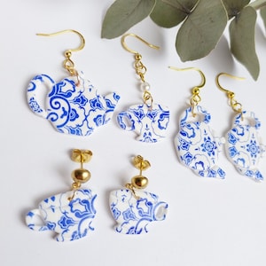 Op de afbeelding: Een set van vier blauwe en witte porseleinen theepot- en theekop-oorbellen. De oorbellen zijn gemaakt van goudkleurig metaal en hebben een delicaat bloemenpatroon.