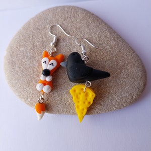 Peut inclure: Une paire de boucles d'oreilles pendantes avec un oiseau noir et un morceau de fromage jaune, et un renard avec une queue blanche. Les boucles d'oreilles sont en pâte polymère et ont des crochets argentés.