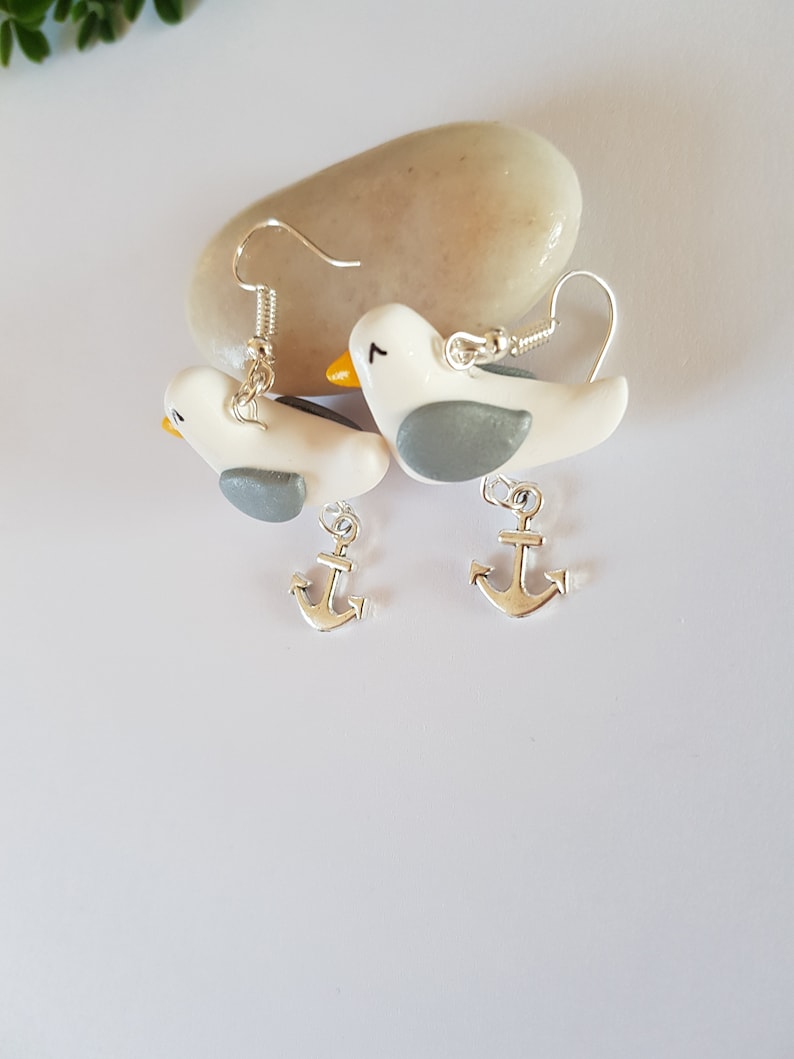 May include: A pair of silver dangle earrings featuring white and gray bird charms with silver anchor charms hanging below.