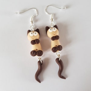 Op de afbeelding: Een paar oorhangers met bruine en beige klei kattenfiguren met witte ogen en bruine staarten.