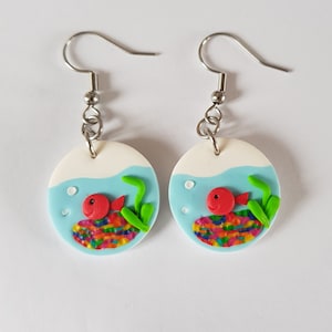 Op de afbeelding: Een paar handgemaakte oorbellen met een grillig aquariumontwerp. Elke oorbel heeft een witte bovenkant, lichtblauw water, een rode vis en kleurrijk grind. Groen zeewier voegt details toe. Zilverkleurige oorhaken.