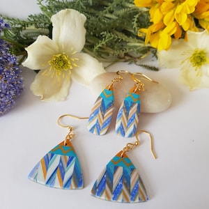 Puede incluir: Un par de pendientes geométricos azules y dorados. Los pendientes están hechos de arcilla polimérica y tienen un patrón de chevron. Los pendientes son de estilo colgante y tienen ganchos dorados.