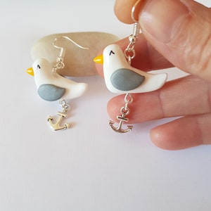 May include: A pair of silver dangle earrings featuring white and gray clay seagull charms with yellow beaks and silver anchor charms.