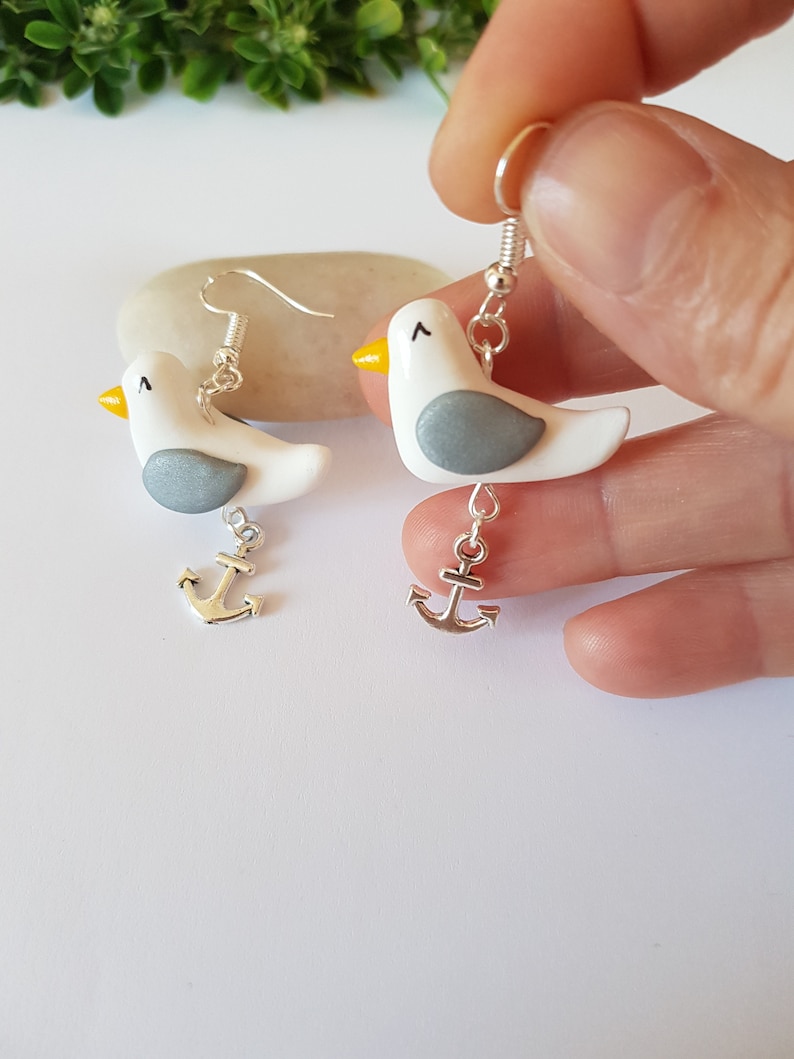 May include: A pair of silver dangle earrings featuring white and gray clay seagull charms with yellow beaks and silver anchor charms.