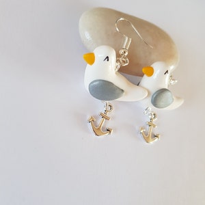 May include: A pair of silver dangle earrings featuring white seagull charms with gray accents and yellow beaks. Each seagull charm is attached to a silver anchor charm.