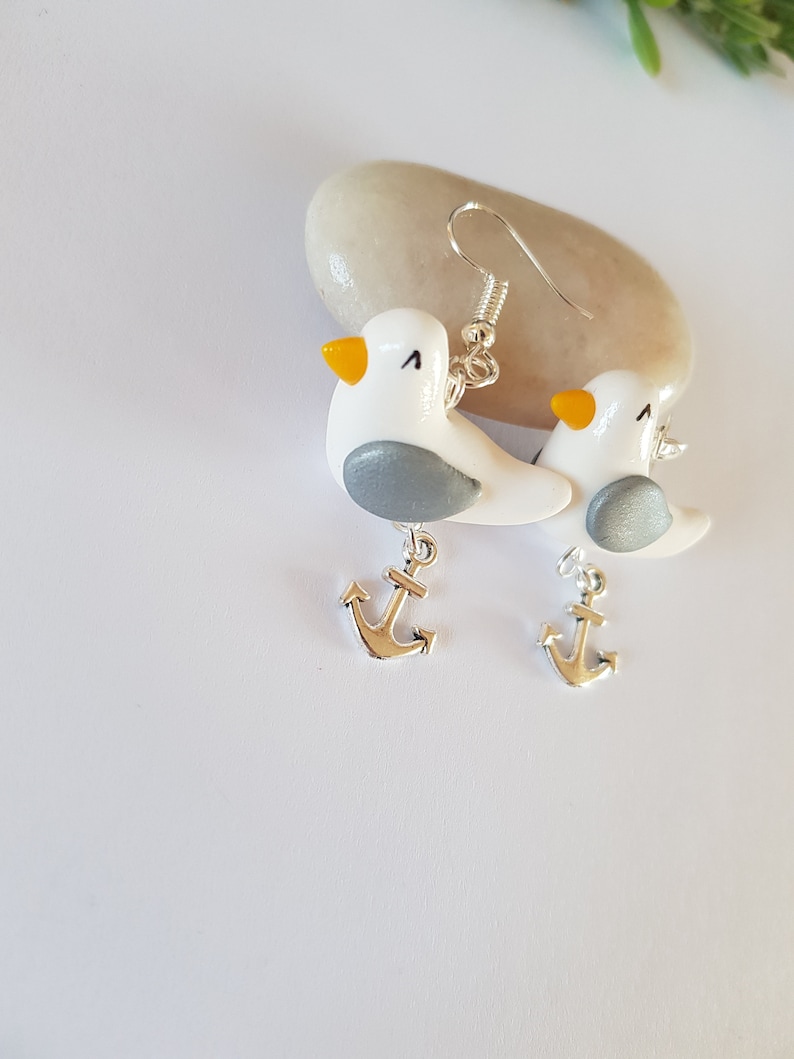 May include: A pair of silver dangle earrings featuring white seagull charms with gray accents and yellow beaks. Each seagull charm is attached to a silver anchor charm.